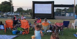 Picture of people sitting at the park on ground and lawn chairs watching movie on inflatable screen